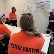 Three traffic controllers are in a room as a Manager is stood up next to a graphic.