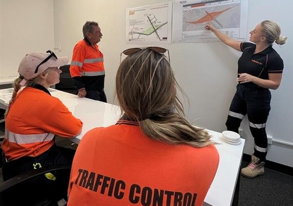 Three traffic controllers are in a room as a Manager is stood up next to a graphic.