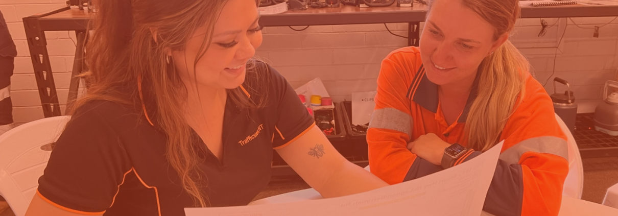 Kayla and Amy sit in the office, looking at paperwork. One girl is in PPE the other is in Black Uniform.