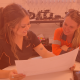 Kayla and Amy sit in the office, looking at paperwork. One girl is in PPE the other is in Black Uniform.